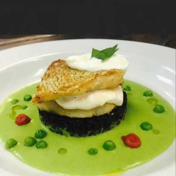 Gourmet dish with stacked fish, white sauce, black rice, green pea puree, and red garnish.