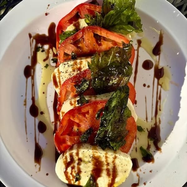 Caprese salad with sliced tomatoes, mozzarella, basil, olive oil, and balsamic glaze on a white plate.